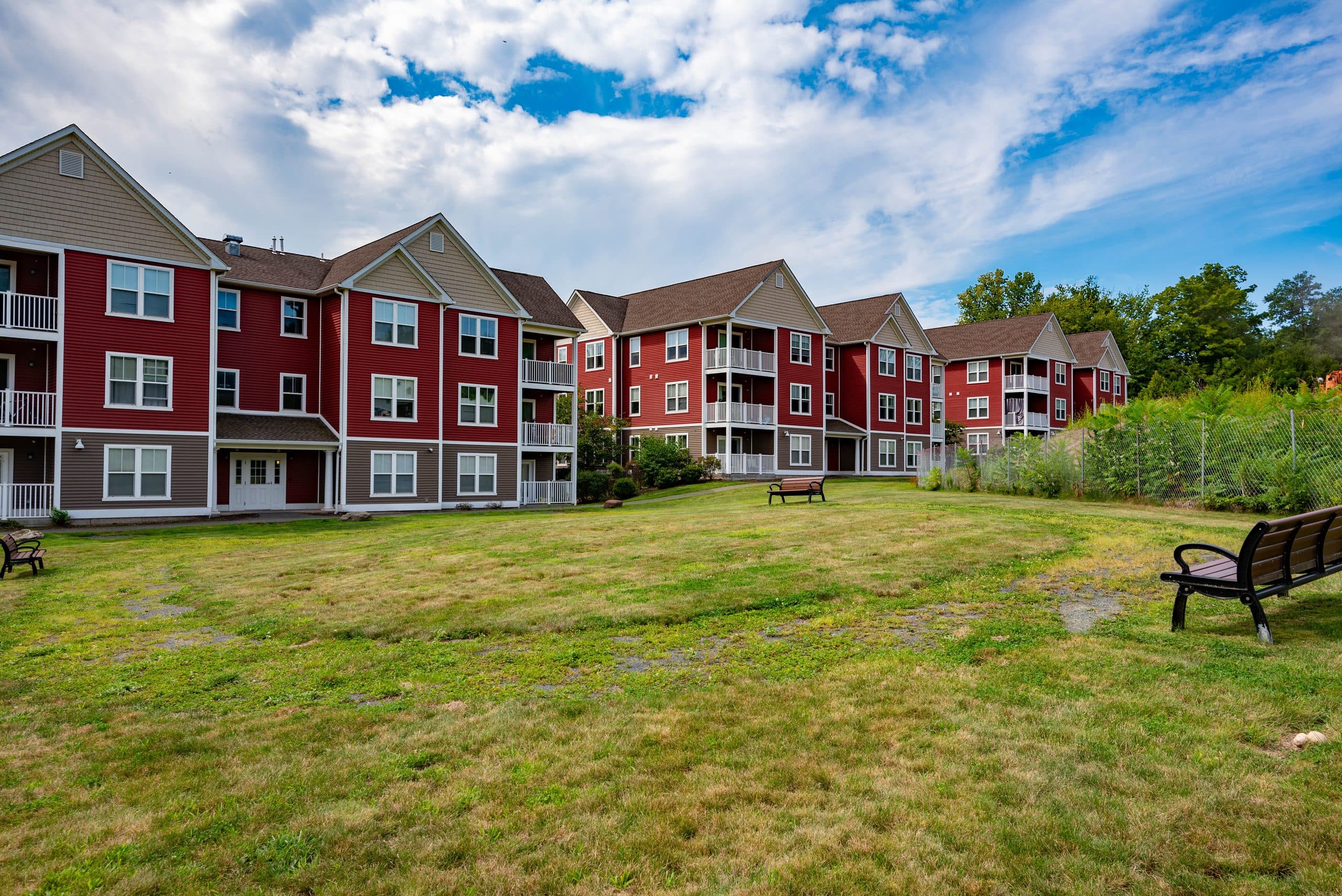 The open green at Brook Hill Village, benches along its edge and the red-clapboard apartment buildings beyond, on a clear summer afternoon