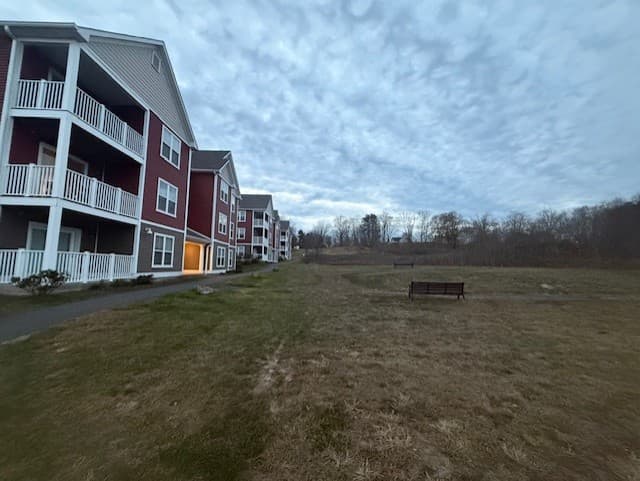 Wide lawn beside the Brook Hill Village buildings with a bench and a tree line beyond