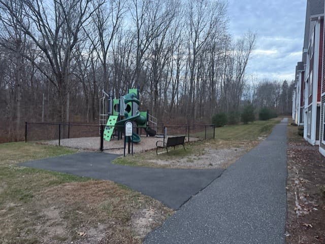 Fenced playground with climbing equipment and a bench beside a paved path at the edge of the woods